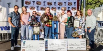 A group stands on a stage holding trophies and large checks in front of a backdrop with Sonoma Marin Fair logos. Four people are holding certificates, celebrating the World's Ugliest Dog 2024 event, with others standing alongside them.