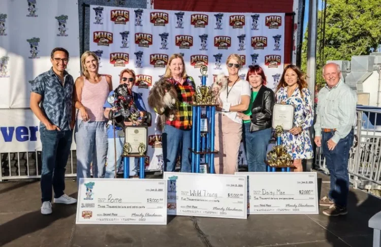 A group stands on a stage holding trophies and large checks in front of a backdrop with Sonoma Marin Fair logos. Four people are holding certificates, celebrating the World's Ugliest Dog 2024 event, with others standing alongside them.