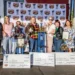 A group stands on a stage holding trophies and large checks in front of a backdrop with Sonoma Marin Fair logos. Four people are holding certificates, celebrating the World's Ugliest Dog 2024 event, with others standing alongside them.