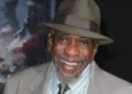 A smiling older man with a beard wearing a green fedora, suit jacket, and striped shirt with a tie resembles Bill Cobbs, an actor known for his remarkable roles in the entertainment industry.