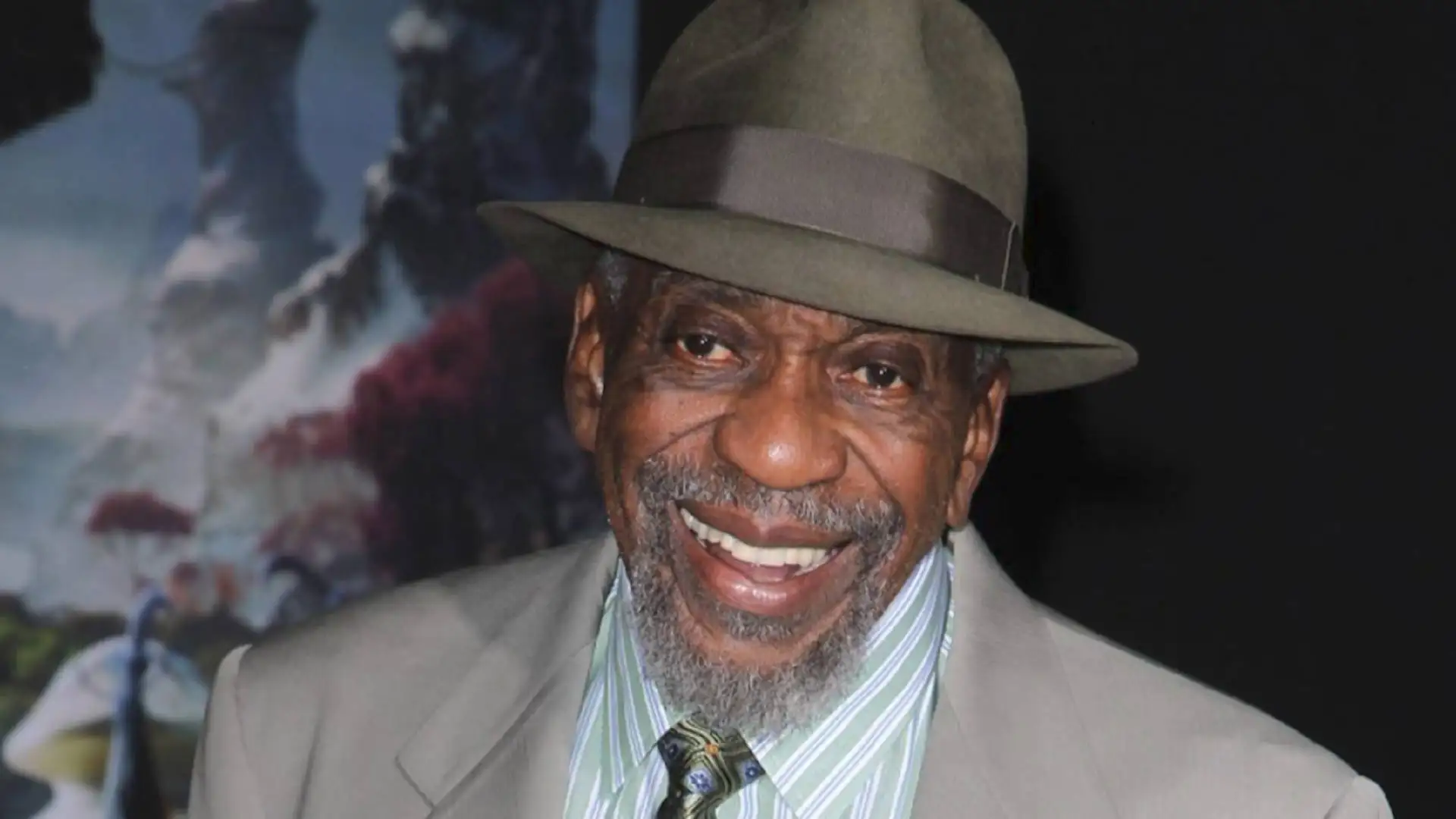 A smiling older man with a beard wearing a green fedora, suit jacket, and striped shirt with a tie resembles Bill Cobbs, an actor known for his remarkable roles in the entertainment industry.