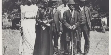 A group of seven people dressed in early 20th century attire stand outdoors, with four men wearing suits and hats, and three women in long dresses and hats, posing for a photo during one of the community events that celebrated Black culture.