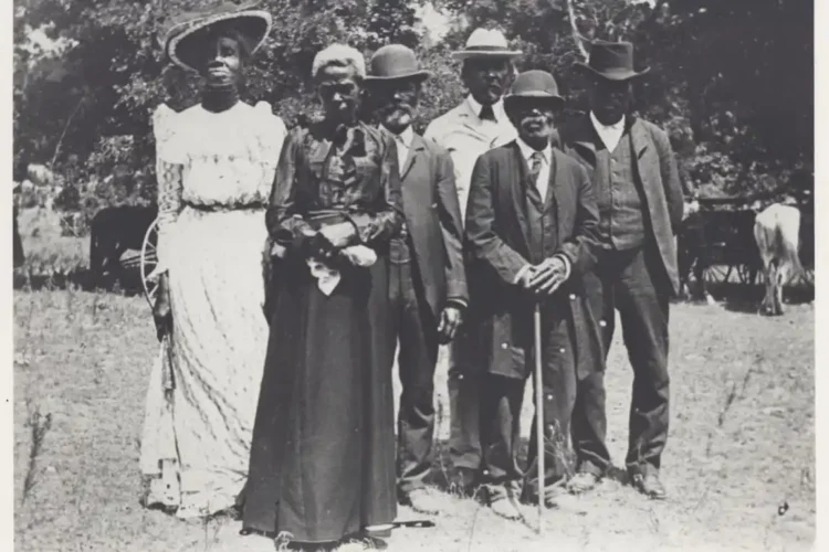 A group of seven people dressed in early 20th century attire stand outdoors, with four men wearing suits and hats, and three women in long dresses and hats, posing for a photo during one of the community events that celebrated Black culture.
