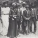 A group of seven people dressed in early 20th century attire stand outdoors, with four men wearing suits and hats, and three women in long dresses and hats, posing for a photo during one of the community events that celebrated Black culture.