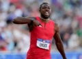 Noah Lyles in a red outfit, "LYLES" on his bib, gestures with his right hand during the 100m Final at the US Olympic Trials, running past a blurred crowd.