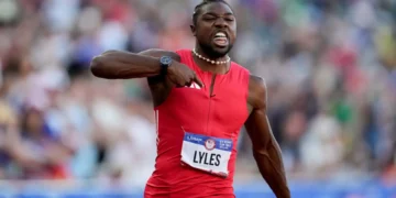 Noah Lyles in a red outfit, "LYLES" on his bib, gestures with his right hand during the 100m Final at the US Olympic Trials, running past a blurred crowd.