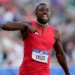 Noah Lyles in a red outfit, "LYLES" on his bib, gestures with his right hand during the 100m Final at the US Olympic Trials, running past a blurred crowd.