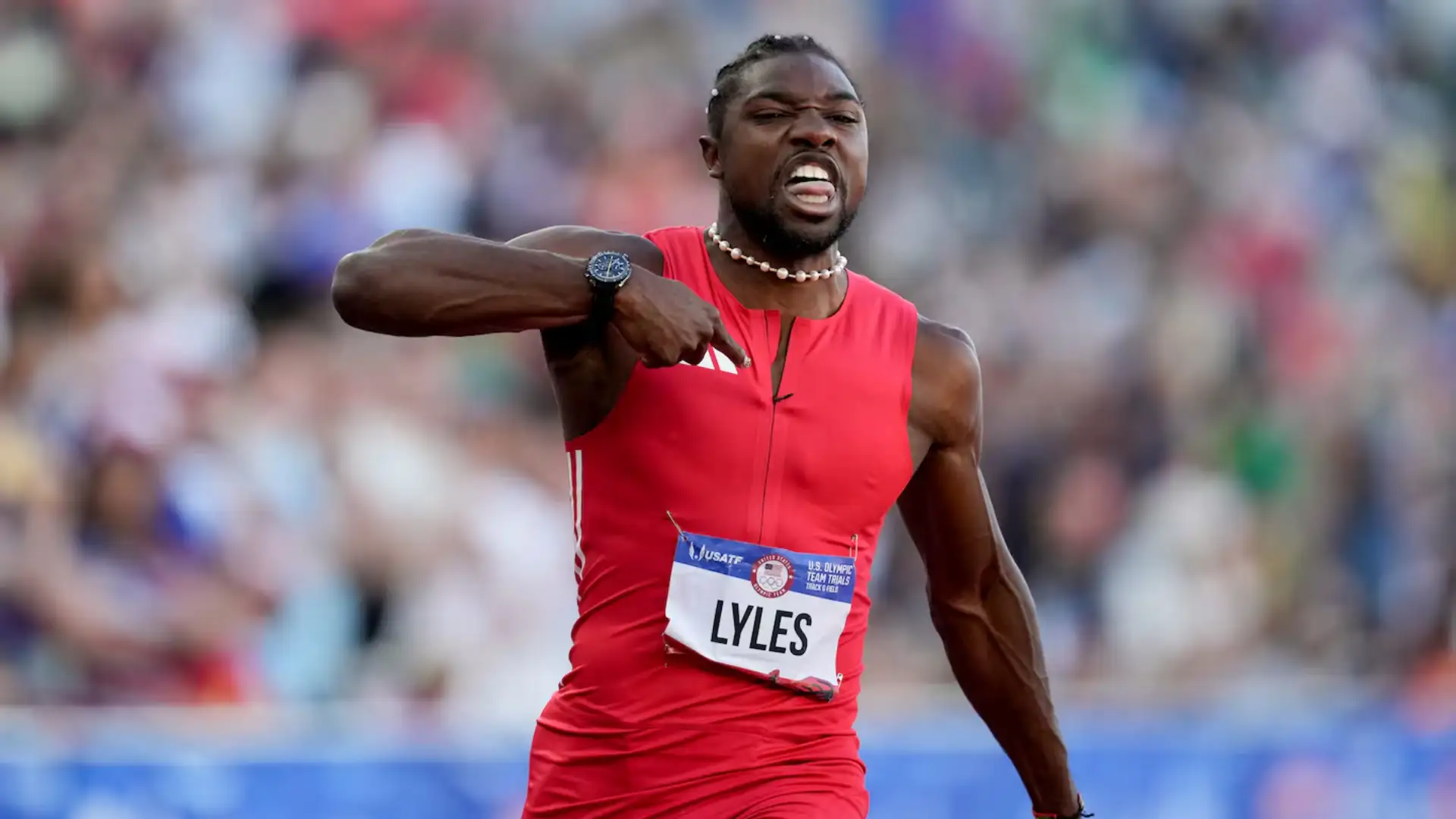 Noah Lyles in a red outfit, "LYLES" on his bib, gestures with his right hand during the 100m Final at the US Olympic Trials, running past a blurred crowd.