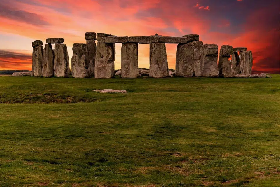Stonehenge monument at sunset, with large standing stones arranged in a circular formation under a vibrant, colorful sky. Nearby, the ongoing Stonehenge road tunnel project has sparked numerous protests.