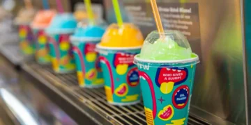 A row of colorful frozen drink cups with dome lids and straws, each containing different brightly colored slush beverages, displayed on a metal rack at 7-Eleven for Free Slurpee Day.