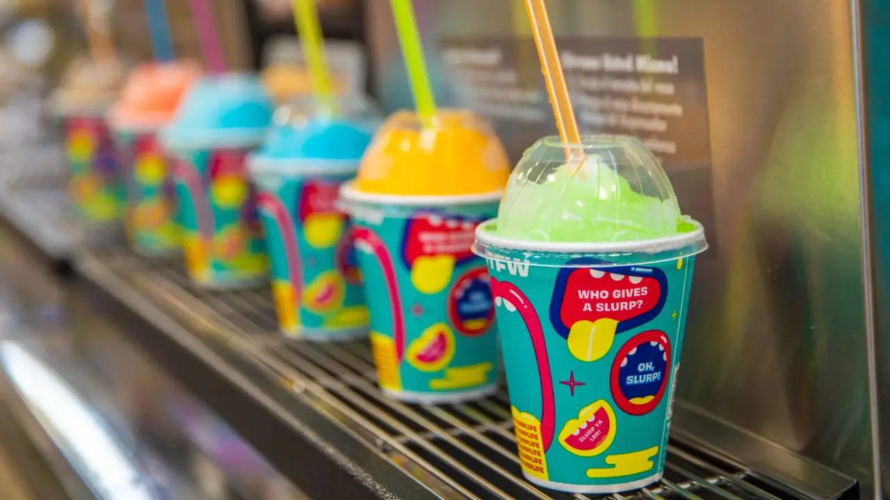 A row of colorful frozen drink cups with dome lids and straws, each containing different brightly colored slush beverages, displayed on a metal rack at 7-Eleven for Free Slurpee Day.