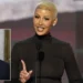 Amber-Rose with a shaved head, resembling Amber Rose, is speaking at a podium. There is a small inset image of another person in a suit and tie in the bottom left corner, reminiscent of the Republican National Convention setup.