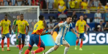 lionel messi in a blue and white striped Argentina jersey runs with the ball while a player in a yellow and blue Colombia jersey attempts to block his path during the Copa América Final 2024, with more players and spectators in the background.