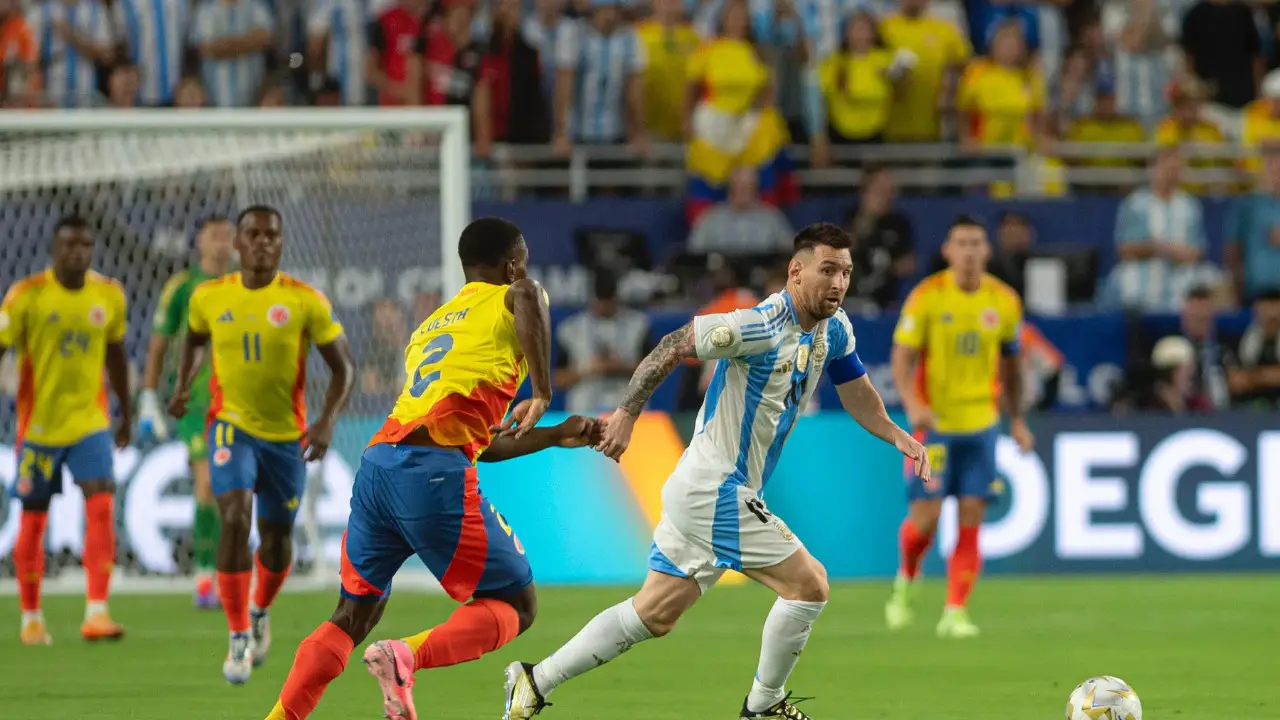 lionel messi in a blue and white striped Argentina jersey runs with the ball while a player in a yellow and blue Colombia jersey attempts to block his path during the Copa América Final 2024, with more players and spectators in the background.