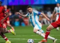 Three soccer players in action: Lionel Messi in a white and blue striped jersey, reminiscent of Lionel Messi in Argentina's Copa America kit, dribbles the ball between two players in red jerseys on a green field.