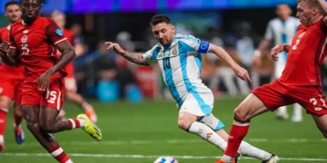 Three soccer players in action: Lionel Messi in a white and blue striped jersey, reminiscent of Lionel Messi in Argentina's Copa America kit, dribbles the ball between two players in red jerseys on a green field.