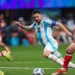 Three soccer players in action: Lionel Messi in a white and blue striped jersey, reminiscent of Lionel Messi in Argentina's Copa America kit, dribbles the ball between two players in red jerseys on a green field.