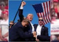Former President Donald Trump in a suit raises his fist while surrounded by security personnel, with an American flag in the background at a Pennsylvania Rally after alleged assassination attempt featuring Former President Donald Trump.