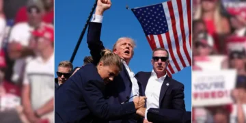 Former President Donald Trump in a suit raises his fist while surrounded by security personnel, with an American flag in the background at a Pennsylvania Rally after alleged assassination attempt featuring Former President Donald Trump.