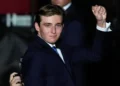 Barron Trump in a navy suit raises his fist in the air at a Florida Rally. He has short, brown hair and a slight smile. There is another person in the background partially visible.