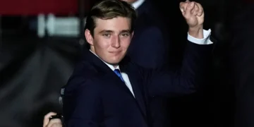 Barron Trump in a navy suit raises his fist in the air at a Florida Rally. He has short, brown hair and a slight smile. There is another person in the background partially visible.