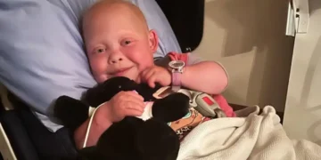 A child with short hair smiles while lying in a hospital bed, holding a black stuffed animal and covered with a white blanket. This inspirational journey belongs to Bella Brave, a young TikTok star who has touched many hearts.