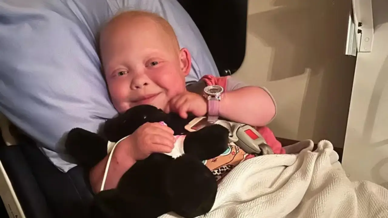 A child with short hair smiles while lying in a hospital bed, holding a black stuffed animal and covered with a white blanket. This inspirational journey belongs to Bella Brave, a young TikTok star who has touched many hearts.
