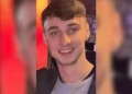 A young man with short brown hair and a gray shirt smiles at the camera against a blurred indoor background with red and yellow tones. The resemblance to missing British teenager Jay Slater, last seen in Tenerife, is striking.