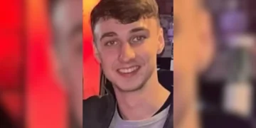 A young man with short brown hair and a gray shirt smiles at the camera against a blurred indoor background with red and yellow tones. The resemblance to missing British teenager Jay Slater, last seen in Tenerife, is striking.