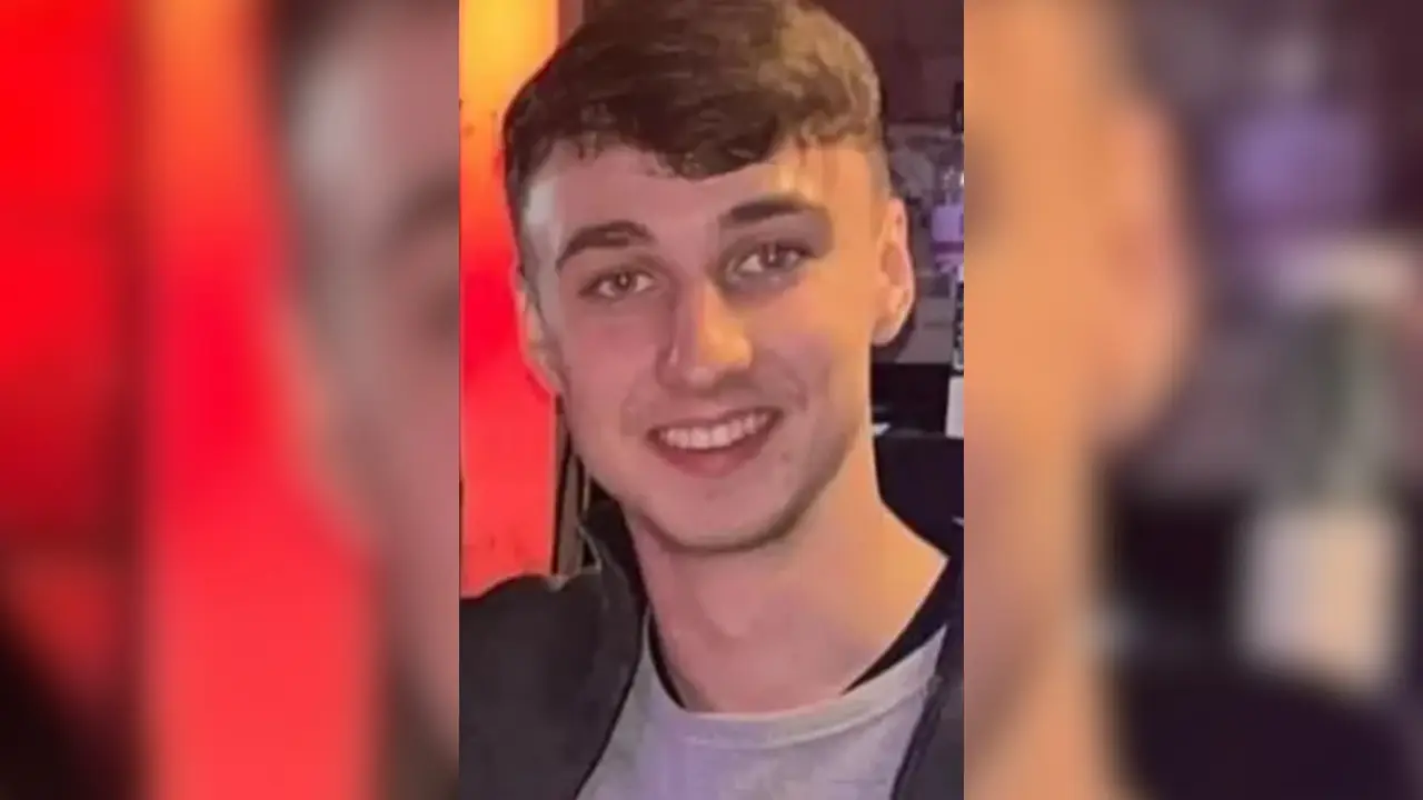 A young man with short brown hair and a gray shirt smiles at the camera against a blurred indoor background with red and yellow tones. The resemblance to missing British teenager Jay Slater, last seen in Tenerife, is striking.