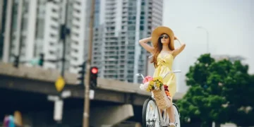 A woman in a yellow dress and sun hat stands beside a white bicycle with a basket of flowers, against an urban backdrop of tall buildings and a bridge. Her choice reflects the trend to reward eco-friendly tourists, making her scene feel straight out of Copenhagen tourism highlights.