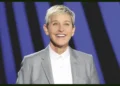 Ellen DeGeneres with short blonde hair wearing a grey blazer and white shirt, smiling against a blue and black striped background, reminiscent of Ellen DeGeneres promoting her Netflix special.