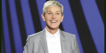 Ellen DeGeneres with short blonde hair wearing a grey blazer and white shirt, smiling against a blue and black striped background, reminiscent of Ellen DeGeneres promoting her Netflix special.