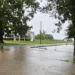 A flooded street in Nashville, IL, with standing water covering the roadway. Trees and buildings are visible in the background under an overcast sky, as officials order evacuation due to a nearby dam failure.