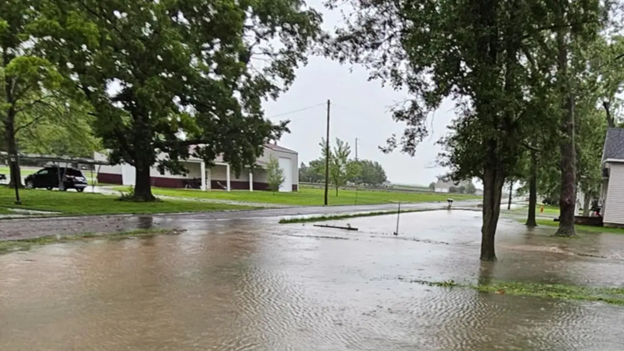 A flooded street in Nashville, IL, with standing water covering the roadway. Trees and buildings are visible in the background under an overcast sky, as officials order evacuation due to a nearby dam failure.