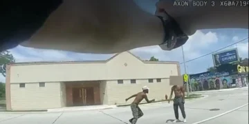 Two individuals in confrontational stances on a Milwaukee street, with a building in the background. A third person's arm, wearing a wristwatch, is visible in the foreground.