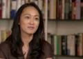 A woman with long, black hair is seated in front of a bookshelf, wearing a brown blouse and a necklace, looking slightly off-camera, as if contemplating matters of national security.
