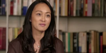 A woman with long, black hair is seated in front of a bookshelf, wearing a brown blouse and a necklace, looking slightly off-camera, as if contemplating matters of national security.