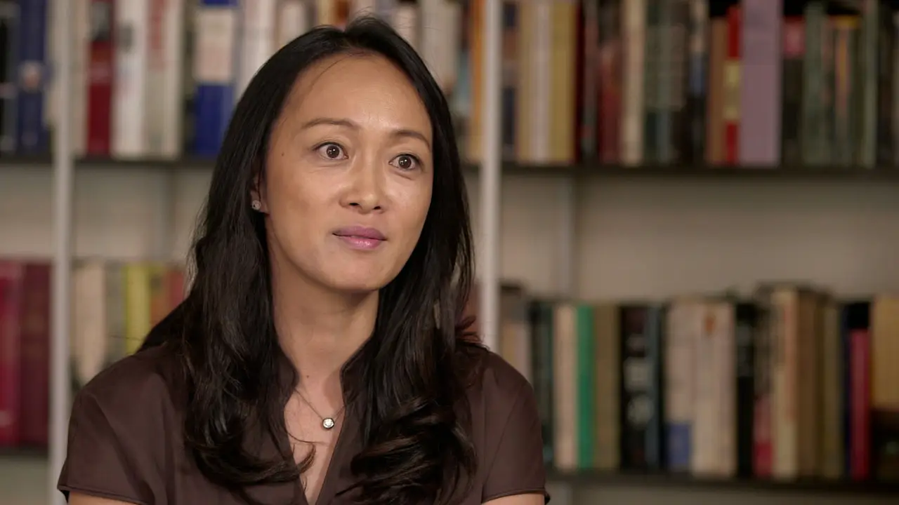 A woman with long, black hair is seated in front of a bookshelf, wearing a brown blouse and a necklace, looking slightly off-camera, as if contemplating matters of national security.