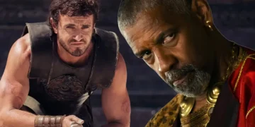 Two men are depicted. The man in the foreground has a serious expression, gray beard, and is wearing a patterned outfit. The man in the background, clad in armor, looks distressed and holds an object in his hands—a scene reminiscent of Gladiator II starring Paul Mescal.