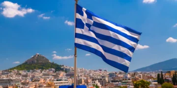 The Greek flag waves proudly on a flagpole against a breathtaking cityscape and mountain backdrop under a clear blue sky, epitomizing the beauty of Greece.
