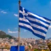 The Greek flag waves proudly on a flagpole against a breathtaking cityscape and mountain backdrop under a clear blue sky, epitomizing the beauty of Greece.
