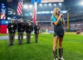 A woman sings the National Anthem into a microphone on a baseball field. She stands in front of a color guard holding flags, delivering a stirring performance despite recent controversy. A large crowd is visible in the stadium.