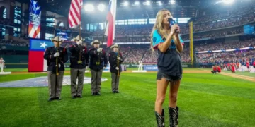 A woman sings the National Anthem into a microphone on a baseball field. She stands in front of a color guard holding flags, delivering a stirring performance despite recent controversy. A large crowd is visible in the stadium.