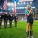 A woman sings the National Anthem into a microphone on a baseball field. She stands in front of a color guard holding flags, delivering a stirring performance despite recent controversy. A large crowd is visible in the stadium.