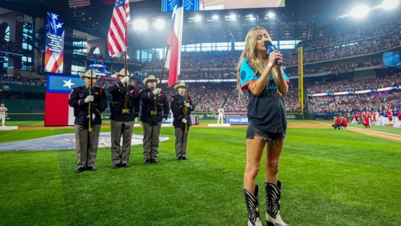 A woman sings the National Anthem into a microphone on a baseball field. She stands in front of a color guard holding flags, delivering a stirring performance despite recent controversy. A large crowd is visible in the stadium.