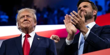Two men dressed in formal suits stand side by side; the man on the left, J.D. Vance, is wearing a red tie and the man on the right is clapping with a neutral expression. Speculation surrounds whether one of them could be Trump’s 2024 Vice Presidential Nominee.