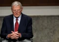 Jim Inhofe wearing glasses, a dark suit, white shirt, and red tie sits with hands clasped, looking forward. The background shows a wooden and concrete interior, reminiscent of an office where Oklahoma Senator Jim Inhofe might discuss the latest legislative matters.
