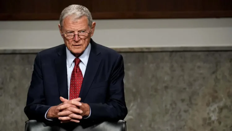 Jim Inhofe wearing glasses, a dark suit, white shirt, and red tie sits with hands clasped, looking forward. The background shows a wooden and concrete interior, reminiscent of an office where Oklahoma Senator Jim Inhofe might discuss the latest legislative matters.