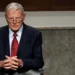 Jim Inhofe wearing glasses, a dark suit, white shirt, and red tie sits with hands clasped, looking forward. The background shows a wooden and concrete interior, reminiscent of an office where Oklahoma Senator Jim Inhofe might discuss the latest legislative matters.